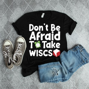 May include: Black t-shirt with white text that reads "Don't Be Afraid To Take WISC-S". A green alarm clock and a red cube are next to the text. The shirt is laid out on a gray surface with a pair of gray sneakers and blue jeans.