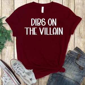 May include: A maroon t-shirt with the text "DIBS ON THE VILLAIN" in white block letters. The shirt is paired with blue jeans and white sneakers. The items are arranged on a wooden surface.