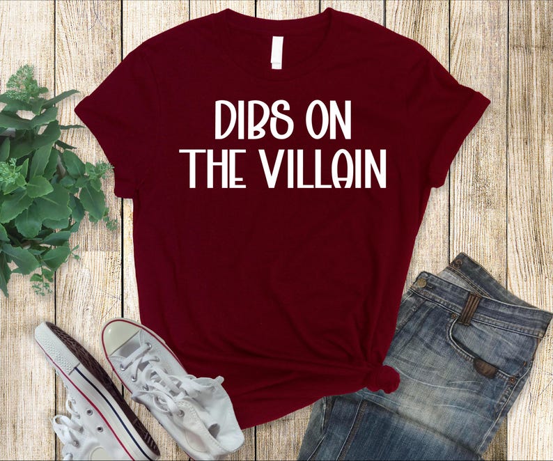 May include: A maroon t-shirt with the text "DIBS ON THE VILLAIN" in white block letters. The shirt is paired with blue jeans and white sneakers. The items are arranged on a wooden surface.