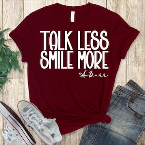 Pu&ograve; includere: Una maglietta bordeaux con la scritta "TALK LESS SMILE MORE" in bianco, con "A Burr" in corsivo sotto. La maglietta &egrave; abbinata a jeans blu e scarpe da ginnastica bianche su uno sfondo di legno.