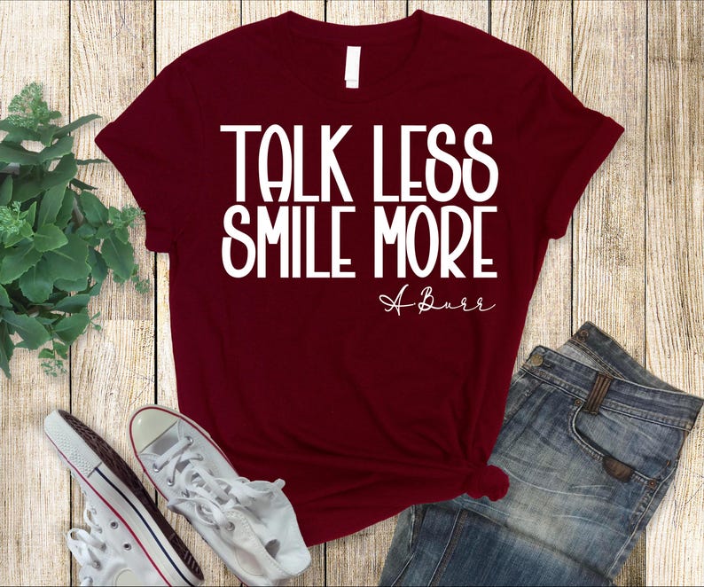 Pu&ograve; includere: Una maglietta bordeaux con la scritta "TALK LESS SMILE MORE" in bianco, con "A Burr" in corsivo sotto. La maglietta &egrave; abbinata a jeans blu e scarpe da ginnastica bianche su uno sfondo di legno.