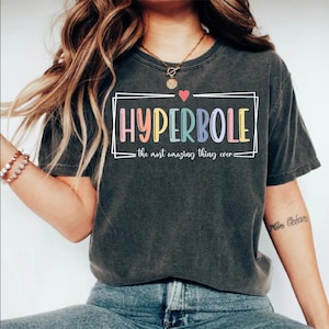 May include: A dark gray t-shirt with a white box around the word "HYPERBOLE" in rainbow colors. The text "the most amazing thing ever" is below the word.