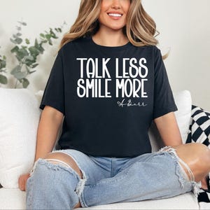 Pu&ograve; includere: T-shirt nero con la scritta bianca "TALK LESS SMILE MORE". La maglietta presenta anche la firma "A. Barr" in corsivo. La modella indossa jeans blu strappati.