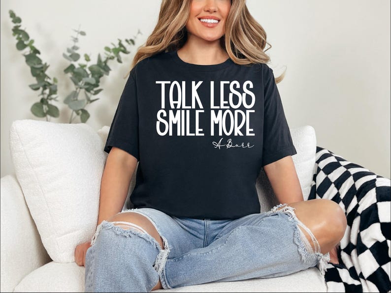 Pu&ograve; includere: T-shirt nero con la scritta bianca "TALK LESS SMILE MORE". La maglietta presenta anche la firma "A. Barr" in corsivo. La modella indossa jeans blu strappati.