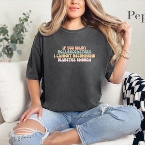 May include: A charcoal gray short-sleeve t-shirt featuring the text "IF YOU ENJOY ROLLERCOASTERS I CANNOT RECOMMEND DIABETES ENOUGH" in a multi-colored retro font. The shirt has a crew neckline and a relaxed fit, with the word "Pepper" visible.