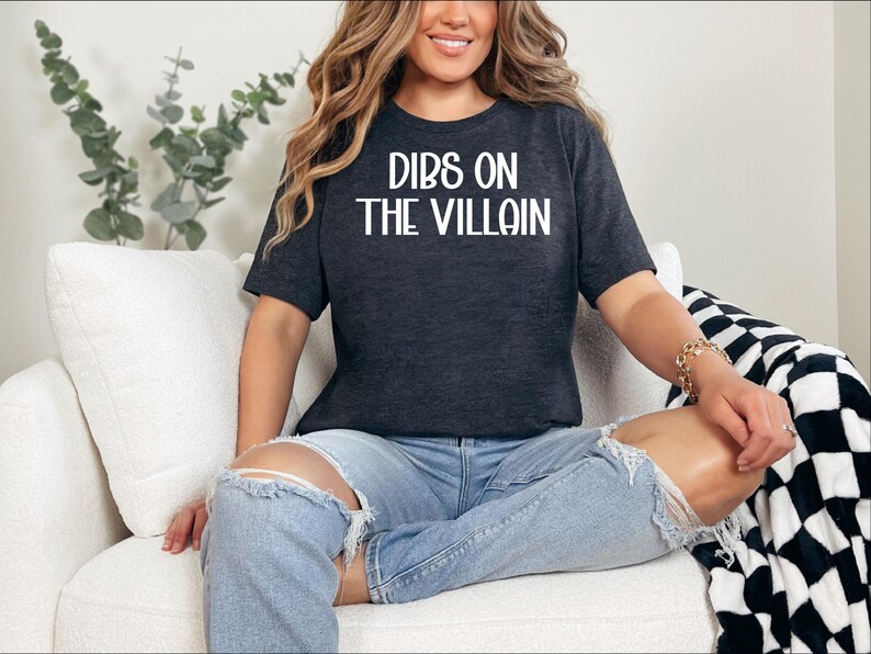 May include: Dark gray t-shirt with the text "DIBS ON THE VILLAIN" in white. The shirt is worn with light blue ripped jeans. The person is sitting on a white couch with a black and white checkered blanket.