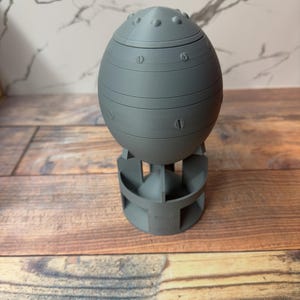 May include: A gray, egg-shaped container with a rounded top and a base. The container has horizontal bands and simulated rivets. The base has a series of vertical supports. The object is made of a solid material.