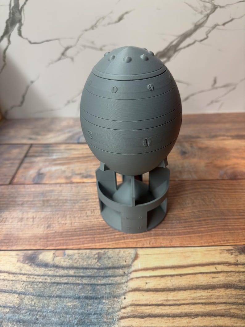 May include: A gray, egg-shaped container with a rounded top and a base. The container has horizontal bands and simulated rivets. The base has a series of vertical supports. The object is made of a solid material.