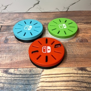 May include: Three colorful game cartridge holders for a Nintendo Switch. The holders are teal, green, and orange, each with a white Nintendo Switch logo. The holders have a black base and a ridged design.