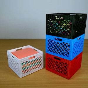 Peut inclure: Trois caisses en plastique colorées empilées, rouge, bleu et noir, avec une petite caisse blanche à gauche. La caisse noire porte le texte "U:A". La caisse blanche contient un objet de couleur corail.