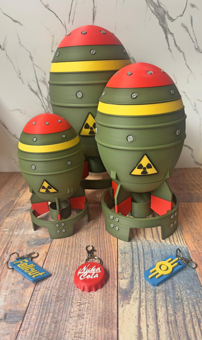 May include: Three olive green, red, and yellow novelty bomb-shaped objects with a radiation symbol, and three keychains. The keychains are blue with "Fallout" text, a red bottle cap with "Nuka Cola" text, and a blue and yellow design.