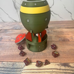 May include: A novelty dice holder shaped like a bomb, in olive green with a yellow band. Red and orange accents are visible on the fins. Several multi-sided dice are scattered around the base, with numbers visible.