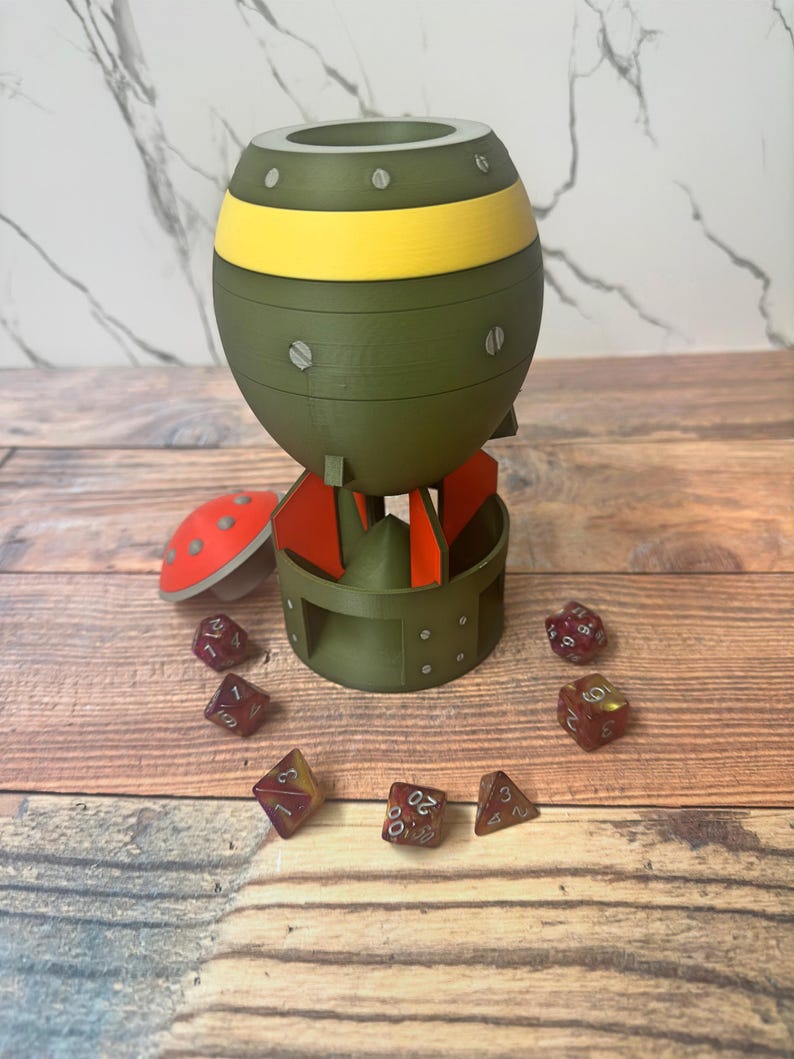 May include: A novelty dice holder shaped like a bomb, in olive green with a yellow band. Red and orange accents are visible on the fins. Several multi-sided dice are scattered around the base, with numbers visible.