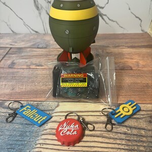 May include: A novelty item set featuring a bomb-shaped figurine with red, yellow, and green accents, along with three keychains. The keychains include a blue Fallout tag, a red Nuka Cola bottle cap, and a blue and yellow emblem. A small parts warning is visible.