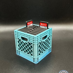 May include: A teal 3D-printed crate-style game cartridge holder with two Nintendo Switch game cartridges. The holder has a lattice design and is designed to hold game cartridges. The cartridges are labeled "Arcade Heat" and "Pokemon".