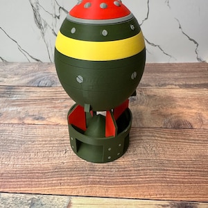 May include: A 3D-printed novelty item shaped like a bomb. The top is red with gray accents, followed by a yellow stripe, and the main body is olive green. It has a circular base with red fins.