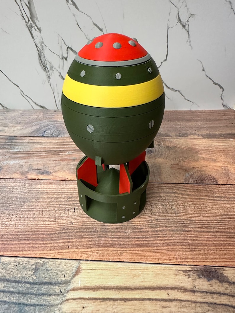 May include: A 3D-printed novelty item shaped like a bomb. The top is red with gray accents, followed by a yellow stripe, and the main body is olive green. It has a circular base with red fins.