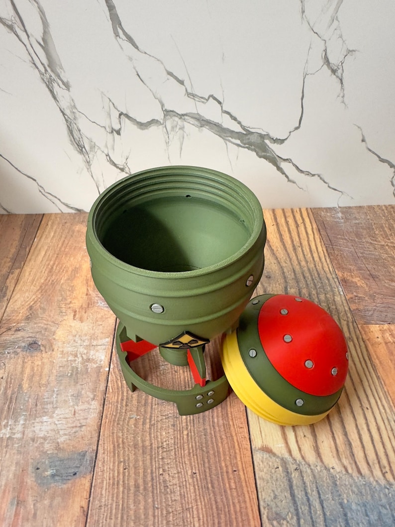 May include: A green, olive-drab colored container with a removable lid. The lid is red with yellow and green accents. The container has a unique, industrial design with visible rivets and a base with red and yellow details.