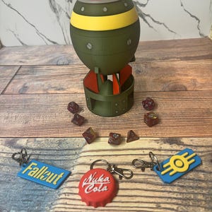 May include: A green, yellow, and red bomb-shaped container with a matching base, surrounded by dice and keychains. The keychains feature the words "Fallout", "Nuka Cola", and a yellow and blue logo.