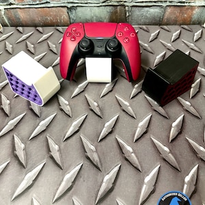 Op de afbeelding: Een rode en zwarte PlayStation 5-controller met een witte standaard en twee controllerhouders. Eén houder is wit met paars interieur, de andere is zwart. De items staan op een traanplaat oppervlak.