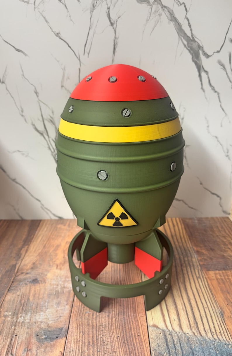 May include: A novelty item shaped like a bomb, featuring a red top, olive green body with a yellow stripe, and a red base. It has a radiation symbol and is made of plastic.