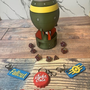 May include: A green, yellow, and red bomb-shaped container with a stand, surrounded by dice and keychains. The keychains are blue and red, with the words "Fallout" and "Nuka Cola" on them. The text "One Free Random Surprise Keychain Included!" is at the bottom.