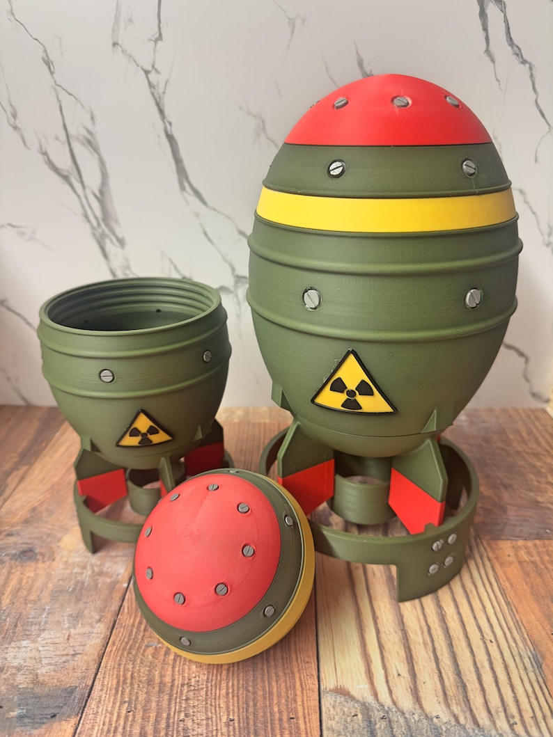 May include: Three-piece set of novelty items resembling miniature bombs. The items are primarily olive green with red and yellow accents. Each piece features a black and yellow radiation symbol. The set includes a container, a bomb-shaped item, and a detached top.