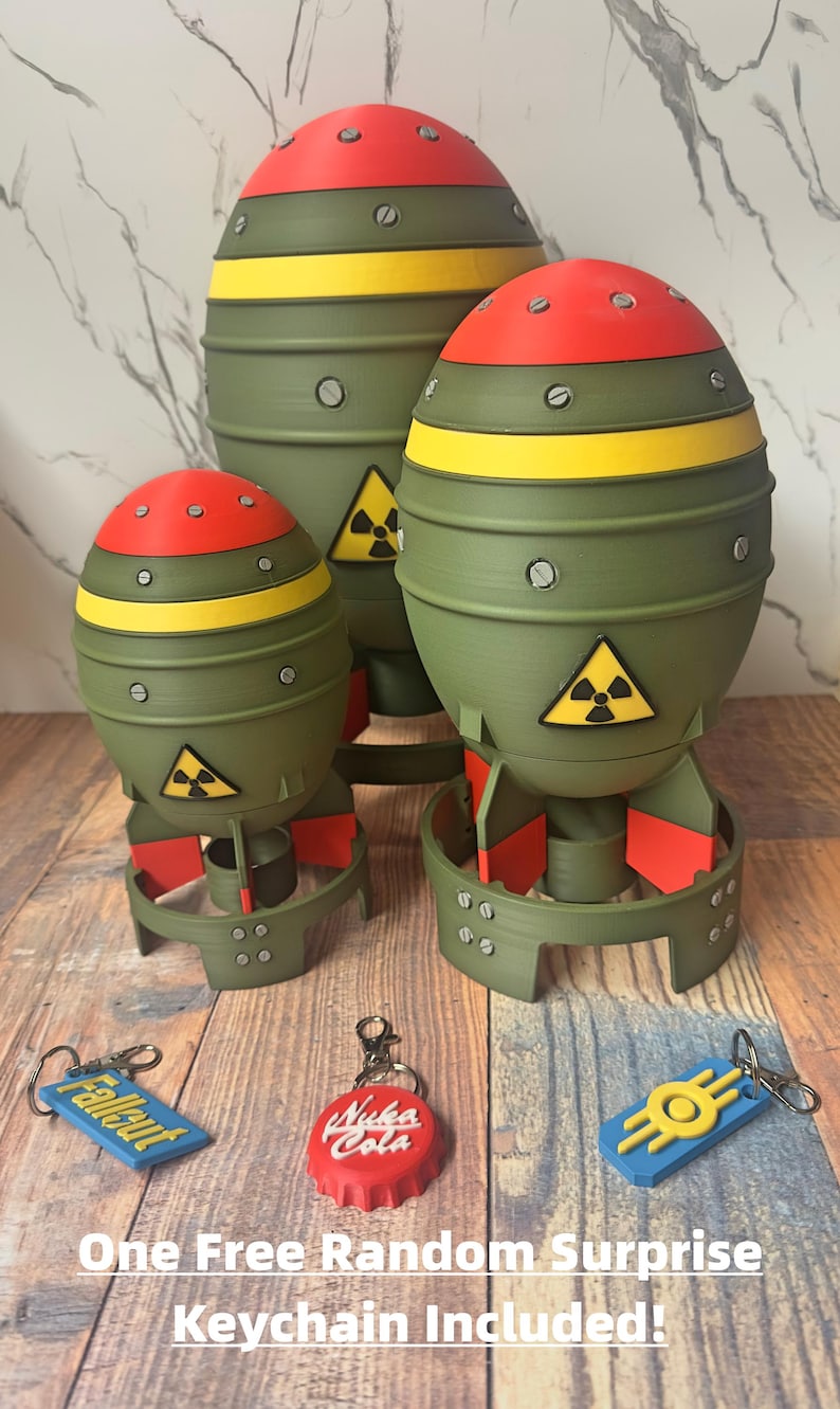 May include: Three olive green, red, and yellow novelty bomb-shaped objects with a radiation symbol. The objects are of varying sizes and sit on a wooden surface. Included are three keychains with the text "Fallout", "Nuka Cola", and a yellow symbol.