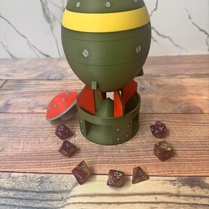 May include: A green and yellow bomb-shaped dice holder with red fins and a red mushroom-shaped lid. Several red and yellow dice are scattered around the holder, all on a wooden surface.