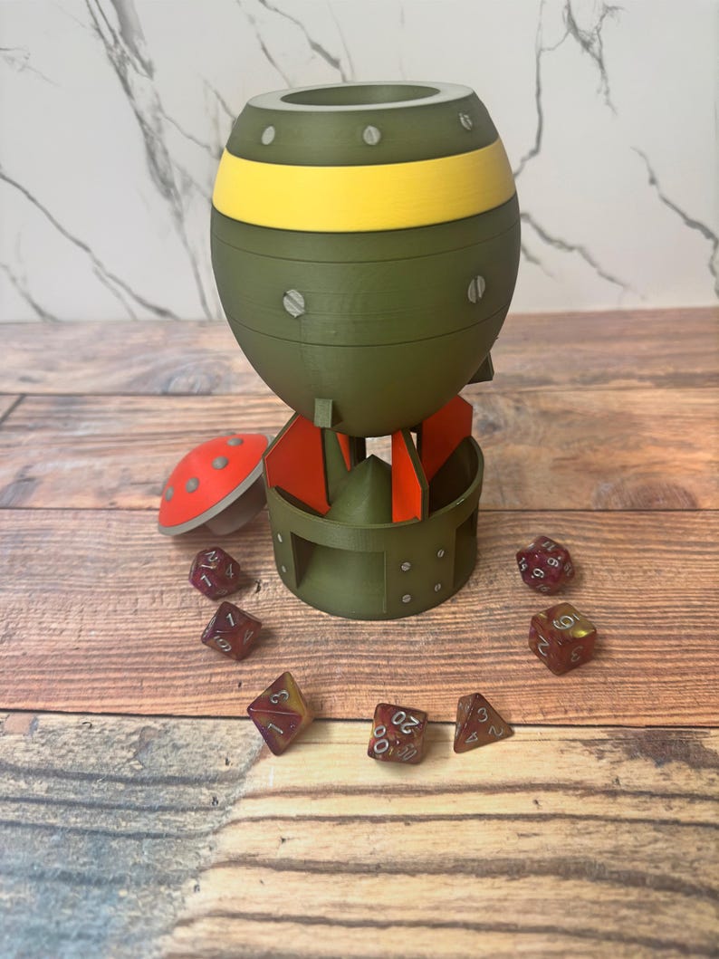May include: A green and yellow bomb-shaped dice holder with red fins and a red mushroom-shaped lid. Several red and yellow dice are scattered around the holder, all on a wooden surface.
