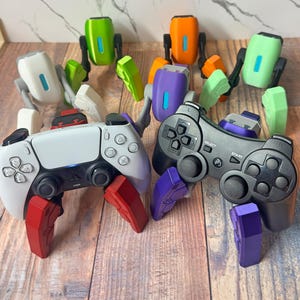 Op de afbeelding: Twee gamecontrollers, één wit en één zwart, met kleurrijke, geometrische opzetstukken. De witte controller heeft rode opzetstukken, terwijl de zwarte controller paarse opzetstukken heeft. Er zijn ook verschillende kleine, robotachtige figuren in verschillende kleuren aanwezig.