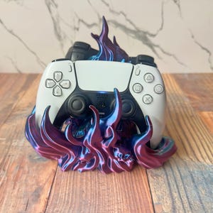 May include: A white and black games controller sits on a decorative stand. The stand features a flame-like design in purple and blue hues. The controller has a symmetrical button layout.