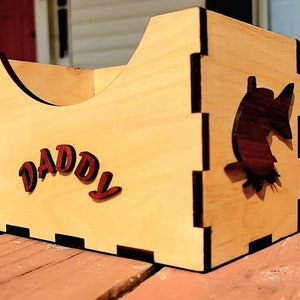 Könnte beinhalten: Eine Holzkiste mit einem Fisch-Ausschnitt an der Seite. Die Kiste ist hellbraun mit einer dunkelbraunen Beize. Das Wort "DADDY" ist in Rot auf der Vorderseite der Kiste geschrieben.
