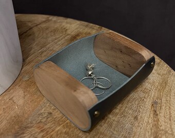 May include: A small grey leather tray with wooden edges, designed to hold jewellery. The tray is sitting on a wooden surface and contains a pair of silver earrings.