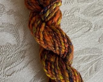 May include: A skein of yarn in shades of orange, yellow, and brown with a hint of purple. The yarn has a sparkly finish and is twisted into a rope shape.