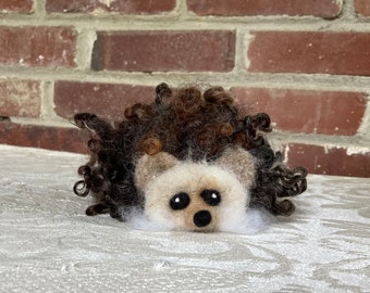 May include: A brown and white needle felted hedgehog with curly brown hair. The hedgehog is sitting on a white surface.