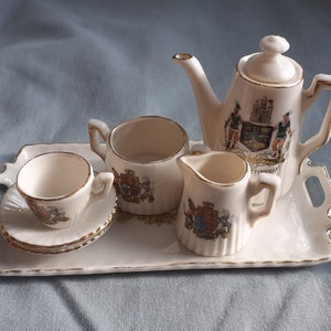 May include: A white porcelain tea set with a gold trim, featuring a coat of arms design. The set includes a teapot, creamer, sugar bowl, and a teacup and saucer. The set is displayed on a white porcelain tray with a gold trim.