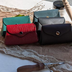 Peut inclure: Un ensemble de quatre petites pochettes en cuir rouge, vert, noir et marron foncé, chacune dotée d'un rabat avec un bouton-pression en métal. La pochette rouge présente une surface texturée, tandis que les autres sont lisses.