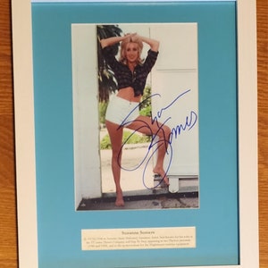 May include: A framed photograph of Suzanne Somers, a blonde woman wearing a white crop top and white shorts, with her signature in blue ink. The text below the photo reads "Suzanne Somers. In 1977-1978 as the iconic 'Chrissy Snow' on ABC's 'Three's Company', Somers has had roles in TV and film, and is the spokesperson for the Thighmaster exercise equipment."