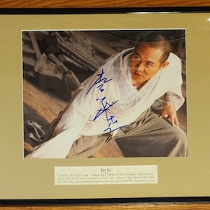 May include: A framed photograph of Jet Li, a Chinese martial artist, actor, and film producer. The photo shows him in a fighting pose, wearing a white shirt and grey pants. The photo is signed by Jet Li with his signature in blue ink. The text below the photo reads "Jet Li, b. 26/4/1963 in Beijing. Chinese Wushu champion, philanthropist, and Actor.  Best known for Once Upon a Time in China, Romeo Must Die, The War, The Forbidden Kingdom, and more films from The Expendables series."