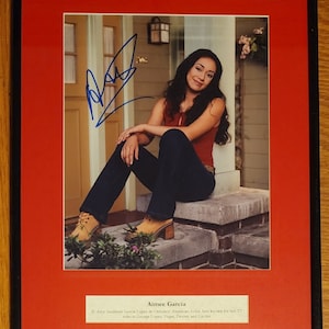May include: Framed photograph of actress Aimee Garcia, signed in blue ink. She is wearing a red top and dark blue jeans, seated on a stone step. The photo is set against a red mat within a black frame. Text at the bottom identifies her.