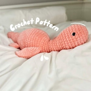 May include: A crocheted pink dinosaur plush toy lying on a white bed. The toy has a long neck and a small head with black eyes. The text "Crochet Pattern" is written in white on a white circle above the toy.