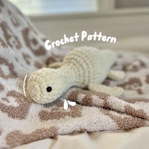 Op de afbeelding: Een wit gehaakt amigurumi dinosaurus speelgoed dat op een bruin en wit deken ligt. Het speelgoed heeft zwarte ogen en een droevige uitdrukking. De tekst "Crochet Pattern" staat boven het speelgoed.