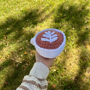May include: A crocheted coffee cup with a white leaf design on a pink background. The cup is being held by a hand wearing a striped sweater.