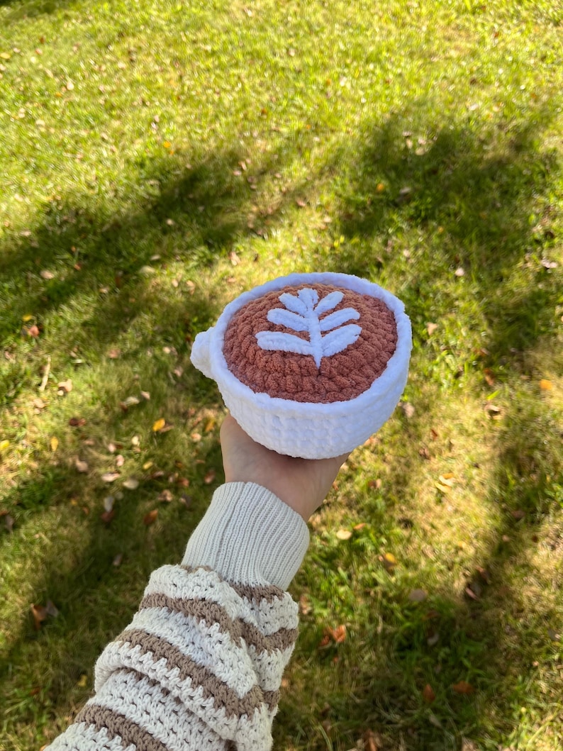 May include: A crocheted coffee cup with a white leaf design on a pink background. The cup is being held by a hand wearing a striped sweater.
