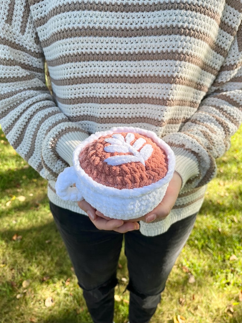 May include: A crocheted coffee cup with a white rim and a pink interior. The cup has a leaf design on the top.