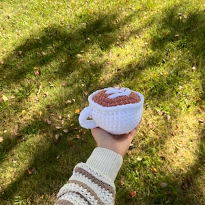 May include: A crocheted coffee cup with a brown and white design. The cup is being held by a hand wearing a striped sweater.