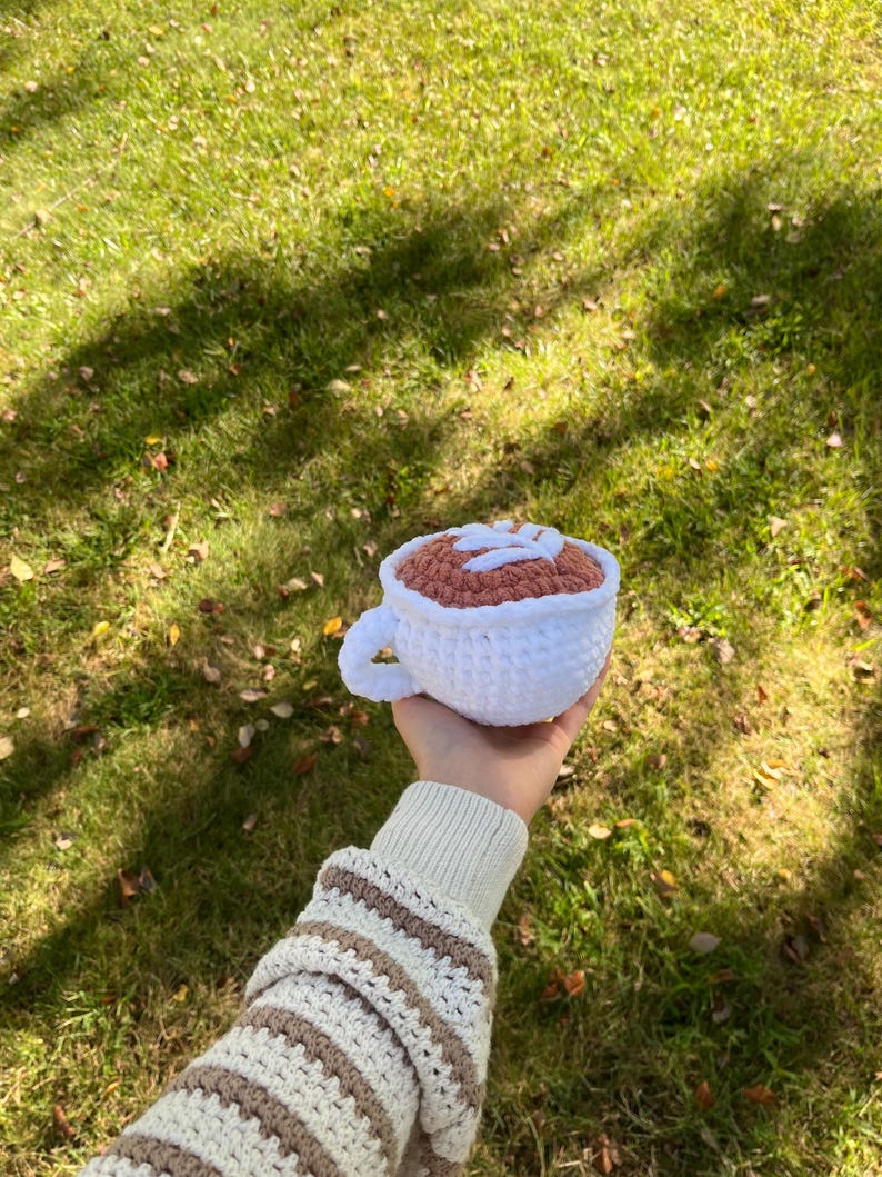 May include: A crocheted coffee cup with a brown and white design. The cup is being held by a hand wearing a striped sweater.