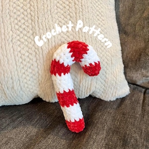 Puede incluir: Un bastón de caramelo tejido a crochet con rayas rojas y blancas. El bastón de caramelo está sobre una superficie blanca y peluda. Crochet Pattern está escrito sobre el bastón de caramelo.