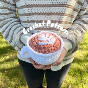 May include: A crocheted coffee cup with a white rim and a pink coffee design. The cup is being held by a person wearing a striped jumper.